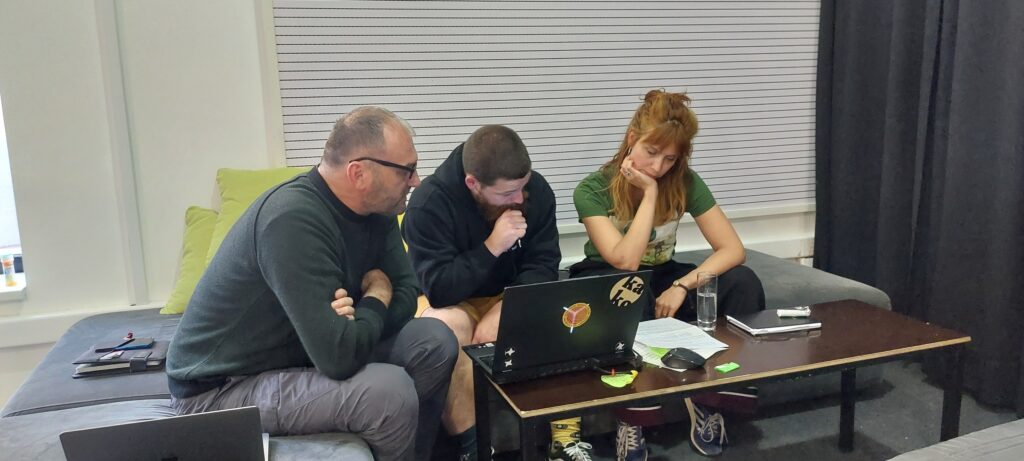 Trije udeleženci sedijo ob mizi in skupaj gledajo v prenosni računalnik.